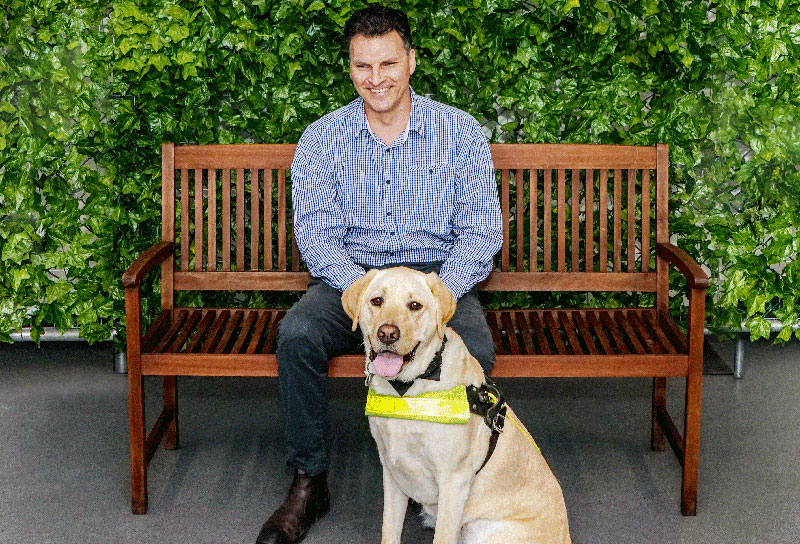 Man sitting on park bench with Guide Dog sitting in front